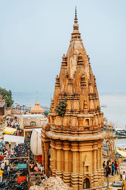 A towering sandstone temple rises on the riverfront of Varanasi, India, surrounded by crowded ghats, boats, and market stalls. The scene captures sacred city life, culture, and spiritual heritage by the Ganges.