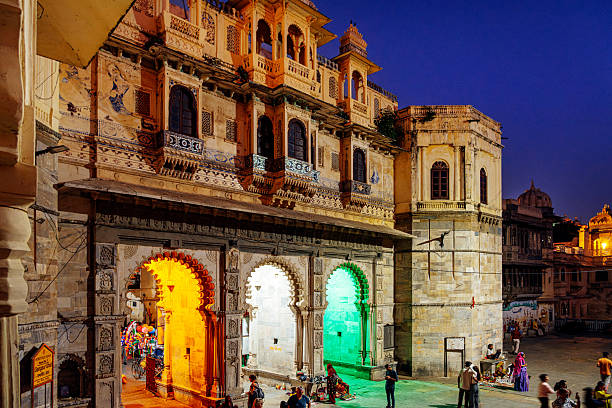 Ornate, illuminated arches form a grand gateway in Udaipur as evening deepens. Warm orange and cool green lights highlight the carved stone, with people and vendors adding life to the heritage scene.