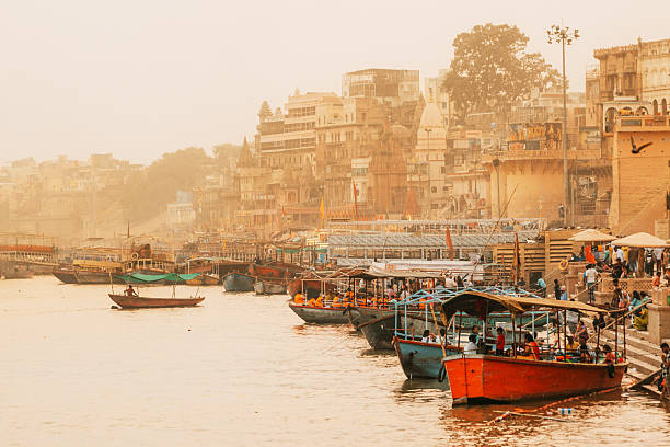 Colorful boats line the Ghats of Varanasi on the Ganges at sunset, with temples and city streets in a hazy backdrop. A vibrant scene of pilgrimage, culture, and river life.