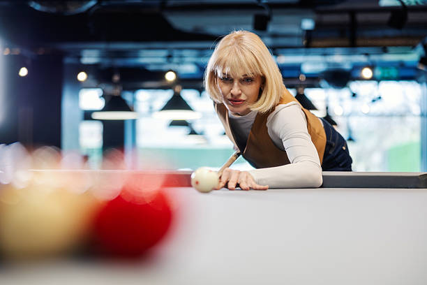 Woman intensely lining up a shot on a pool table with cue stick and cue ball, focused and confident during a casual game in a lively indoor bar setting