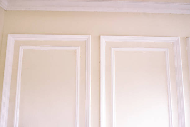 Close-up of an elegant interior wall featuring white crown molding and decorative panel trim. The light cream colored wall provides a classic and sophisticated background for design projects.