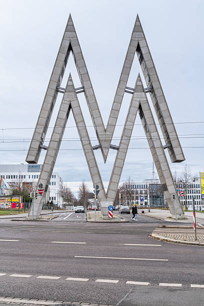 A striking M-shaped sculpture in Leipzig, Germany, blending modern architecture with urban life. Pedestrians and cars emphasize scale and the city’s contemporary public art.