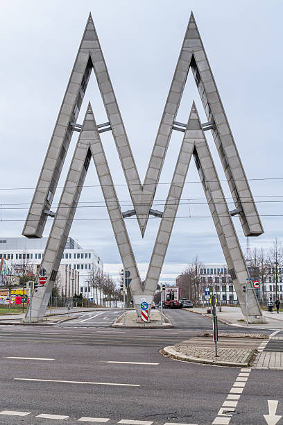 A striking M-shaped steel sculpture stands at a roundabout in Leipzig, Germany. The bold, geometric structure dominates the urban landscape, blending modern art with city infrastructure and creating a memorable landmark.