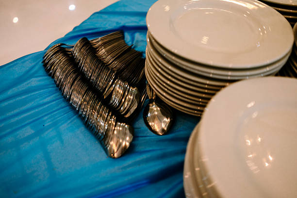 Stacks of pristine white plates and rows of shiny metal spoons are neatly arranged on a vibrant blue tablecloth, prepared for a large gathering or catering event.