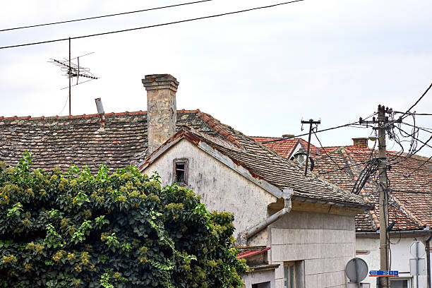 An old rural house with weathered walls, rusty roof edges and a tiny broken window stands beside dense green ivy under a cloudy sky, capturing quiet decay and timeless village charm