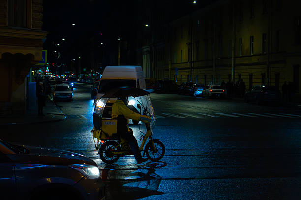 Saint-Petersburg, Russia - 1 November 2025. Person rides electric bicycle with transparent rain canopy on wet street. Cars, van, buildings, streetlights visible