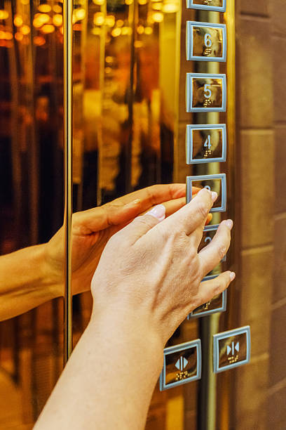 A hand is reaching out to press an elevator button in a modern building with bright lights.