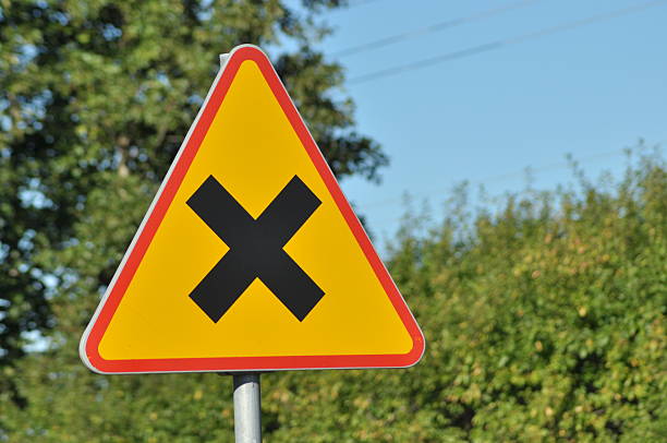 A triangular warning sign with a black cross stands tall beside a road. Lush green trees can be seen in the background under a clear blue sky during daytime.