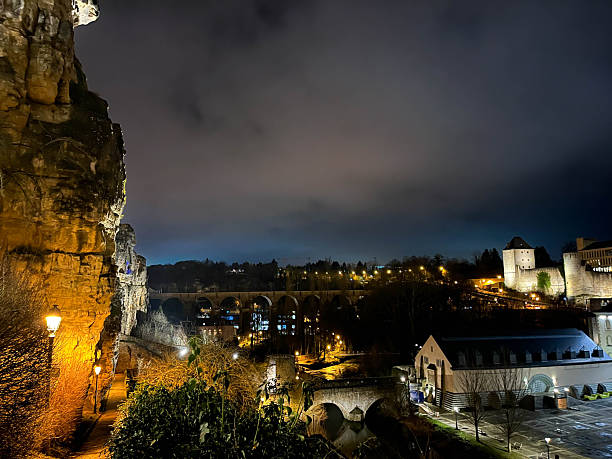 Night view of a steep rocky cliff beside an empty cobblestone path overlooking a softly illuminated valley in Luxembourg City, creating a dramatic, quiet, and atmospheric urban landscape.