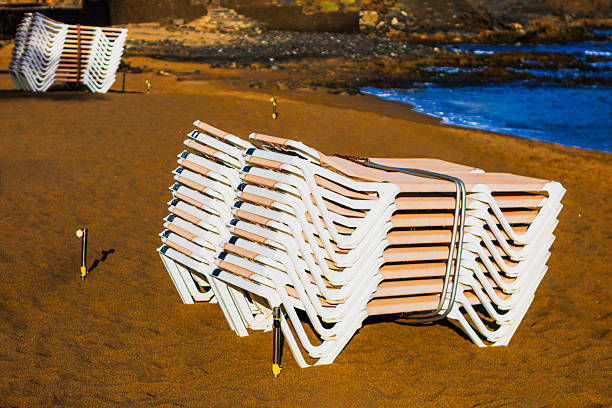 Stacks of white plastic lounge chairs rest on quiet sandy beach, neatly arranged and strapped. Empty shoreline and gentle waves evoke a calm atmosphere awaiting visitors