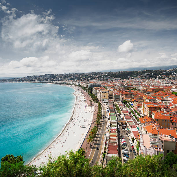 Elevated panoramic view of the curving coastline and Promenade des Anglais in Nice, France, with a crowded sandy beach, turquoise Mediterranean Sea, and dense cityscape along the waterfront. A treetop frames the foreground under a bright, partly cloudy sky, leaving copy space over the water. Unrecognizable tourists appear as small figures on the beach and promenade, conveying a sunny springtime or summer holiday atmosphere.