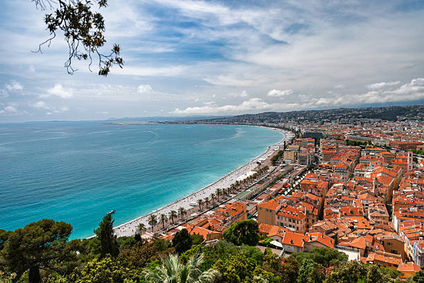 Elevated panoramic view of the curving coastline and Promenade des Anglais in Nice, France, with a crowded sandy beach, turquoise Mediterranean Sea, and dense cityscape along the waterfront. A treetop frames the foreground under a bright, partly cloudy sky, leaving copy space over the water. Unrecognizable tourists appear as small figures on the beach and promenade, conveying a sunny springtime or summer holiday atmosphere.