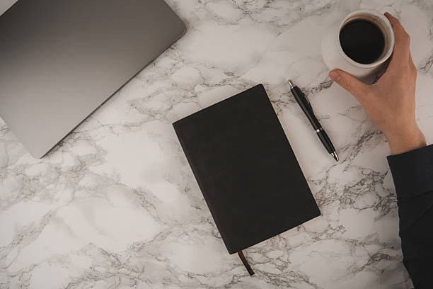 A hand holds a coffee cup while a black notebook and pen lie next to a laptop on a marble table. This scene captures a calm morning workspace atmosphere perfect for productivity