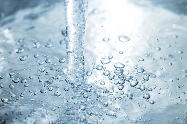 A macro shot of a steady water stream entering a clear liquid, creating bubbles and ripples. Cool blue tones evoke freshness, cleanliness, and motion for dynamic stock content.