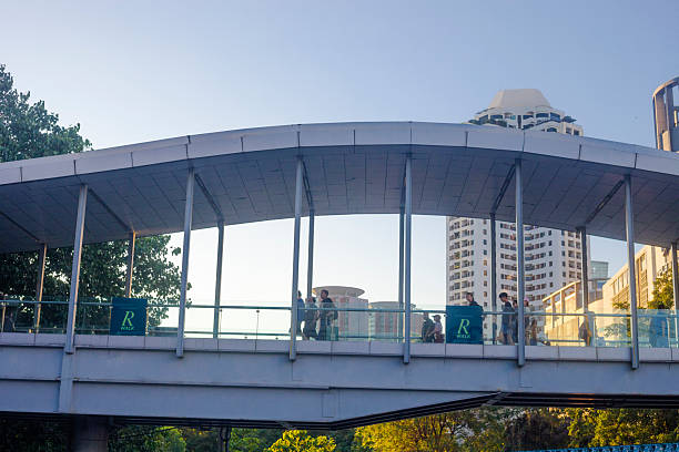 People are walking on R Walk in Bangkok - footbridge is also called Ratchaprasong SkyWalk. Skywalk is in Ratchadamri Road and is leading to shopping mall Central World. Scene is near Pathum Wananurak Park Late afternoon shot. In background are skyscrapers