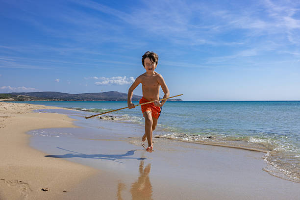 Cheerful little boy running on the beach with a bamboo stick. Summer holiday, fun, and childhood happiness. Jumping boy, beach play, natural setting, candid and authentic feel.