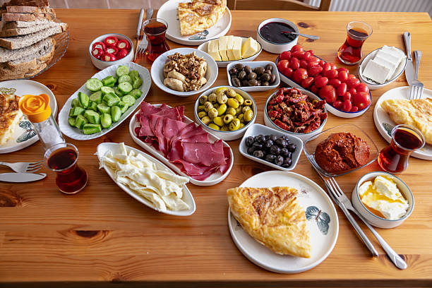 A hearty overhead shot showcasing the abundance and variety of a traditional Turkish spread breakfast. A rich tableau is set on a wooden table, symbolizing hospitality and sharing. Ceramic bowls containing fresh parsley and sliced ​​tomatoes, open plates laden with honey, clotted cream, and various homemade jams (cherry, fig, strawberry) are noteworthy. Fresh white cheese, aged kashar cheese, various olives, and boiled eggs complete the spread. Warm simit (sesame bread rings) and lavash bread are served with roasted peppers and sliced ​​cucumbers. In the background, steaming teacups from the teapot evoke the tradition of Turkish tea. The composition reflects the warm atmosphere of a pleasant Sunday morning spent with family and friends in an intimate kitchen setting.