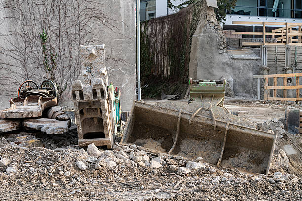 Heavy equipment and debris are scattered at a construction site. Broken machinery and dirt cover the area. Surrounding structures show signs of demolition. Workers may be preparing for new builds.