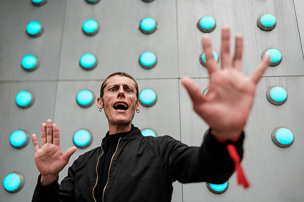 Intense man with wide eyes and open mouth shouting, using both hands in a stop gesture, conveying panic and fear in front of a modern wall with glowing blue industrial lights
