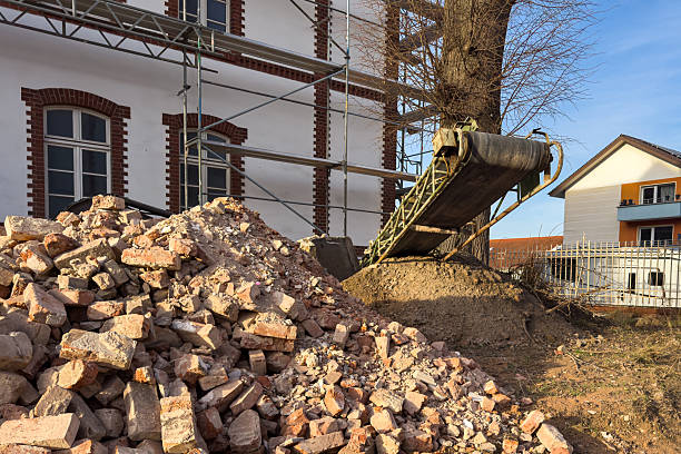 Workers are at a construction site where bricks are piled up near a building. Scaffolding is set up and a conveyor belt is moving materials. It is daytime and the weather looks clear.