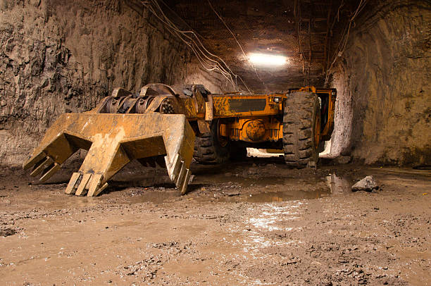 A massive yellow mining machine used to crush rocks stands in a rugged mine tunnel surrounded by damp mud and stone walls. The heavy equipment signals the ongoing mining and industrial operations in the underground mining environment.
