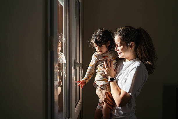 Mother holding her young child, a toddler, looking out a window together as warm morning sunlight streams into their home