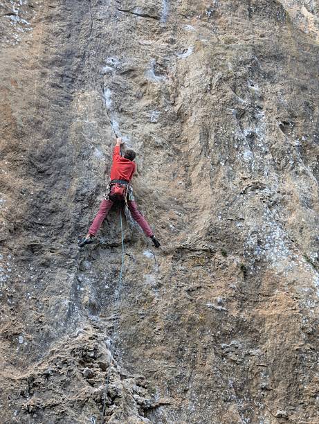 A man wearing a helmet and a blue jacket with an orange vest clips in a rope while climbing a rocky outdoor wall. The focused climber demonstrates rock climbing technique and safety.