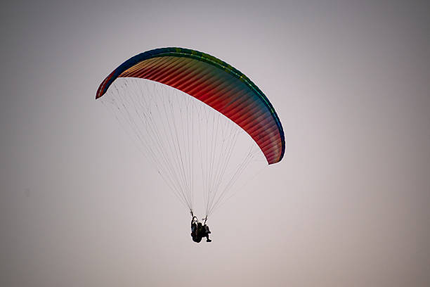 A wide-angle shot of a person paragliding with a multi-colored wing against a hazy coastal background, symbolizing freedom and exploration.