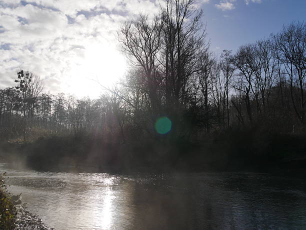 This landscape photograph shows a section of the Wurm River in winter. A light mist hangs over the small river. The low sun is reflected in the water. In the background, the silhouettes of bare trees and a sky with a few clouds are visible.The photo was taken in the Wurm Valley near Herzogenrath (North Rhine-Westphalia, Germany).