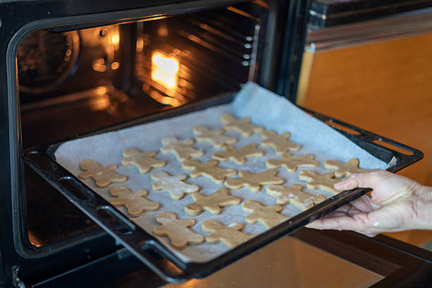 Person's hands carefully placing a baking tray filled with raw gingerbread men cookies on parchment paper into a preheated oven, preparing for holiday baking