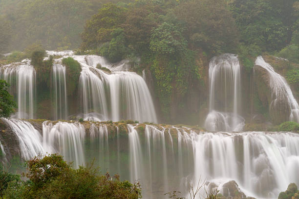 Ban Gioc Waterfall pours over broad, tiered rock ledges into a calm pool, surrounded by dense forest and rising mist. Multiple curtain-like streams descend across the cliff face in a humid natural setting.
