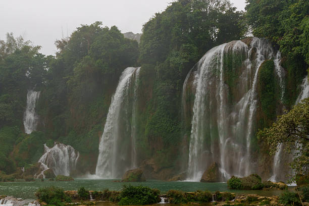 Curtain-like streams flow over limestone cliffs along the side of Ban Gioc Waterfall, descending into a turquoise pool below.