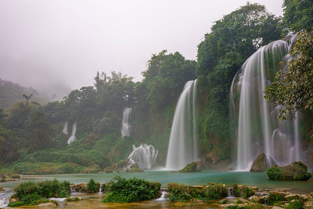 Side cascades of Ban Gioc Waterfall descend over moss-covered limestone cliffs into a calm turquoise pool. Dense forest and mist surround the waterfall sections along the cliff face.