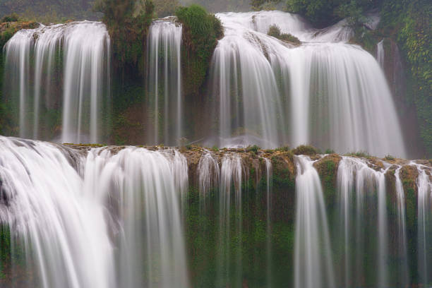 The main curtains of Ban Gioc Waterfall flow over tiered limestone cliffs in smooth, vertical streams. Mist softens the rock face and surrounding vegetation along the waterfall tiers.