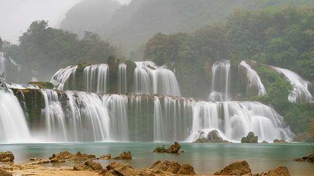 Ban Gioc Waterfall flows in broad, tiered curtains over limestone cliffs into a calm turquoise pool, with rocks and sandy banks in the foreground. Mist hangs over the forested hills surrounding the waterfall.