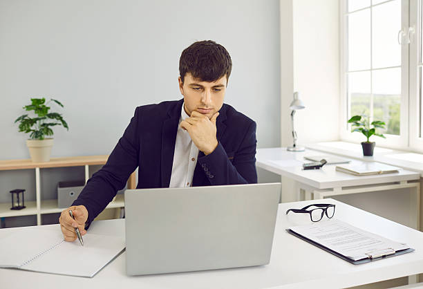 istock Man sitting at office desk with business documents, looking at laptop computer and thinking 2254556815