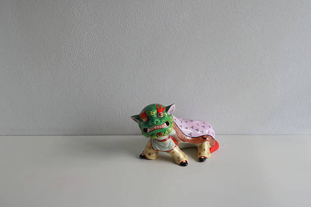 Multicolored Chinese lion dance statue placed on white wooden table against white concrete wall. The Chinese lion dance is a traditional performance art in which two dancers operate a large, colorful lion costume to mimic a lion's movements. The two performers, one controls the head, eyes, ears, and mouth, while the other forms the tail and hind legs. This practice is believed to bring good luck and fortune while driving away evil spirits, and is a staple at the Chinese New Year, business openings, weddings, and other major cultural celebrations.