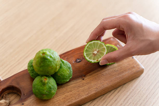 Hand holding a sliced keffir lime above whole limes on wooden cutting board.
