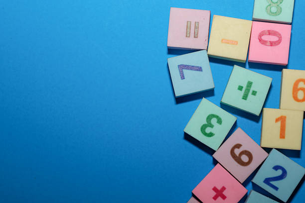istock ​A vibrant educational composition of multicolored toy blocks featuring numbers and arithmetic signs, set against a rich blue background. The pastel-colored cubes (pink, yellow, green, light blue) are grouped on the right side of the frame, 2254578807