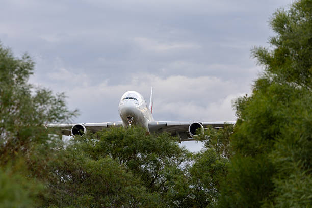 Emirates Airbus A380 wide-body passenger aircraft in flight with landing gear deployed, viewed from a low angle against a backdrop of overcast skies. The double-decker jet features the airline's distinctive white fuselage accented with red and green Emirates branding on the tail and body. Four engines are visible under the wings, and the plane appears to be on final approach or take-off. No people are visible in the image. The setting is outdoors in an aviation context, with neutral grey clouds dominating the scene, suggesting daytime conditions. Main focal points include the aircraft's aerodynamic design, landing gear struts, and winglets. Colours are primarily white, red, green, and grey tones from the sky.