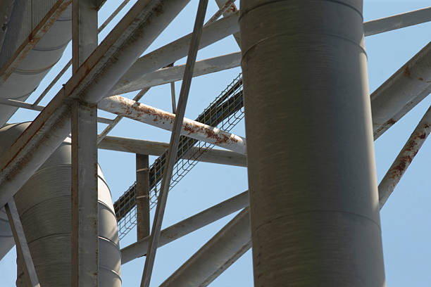 vertical support column meets truss intersection, heavy steel pillar and beams forming load path with visible joints and fasteners, textured surface and sky backdrop suggest