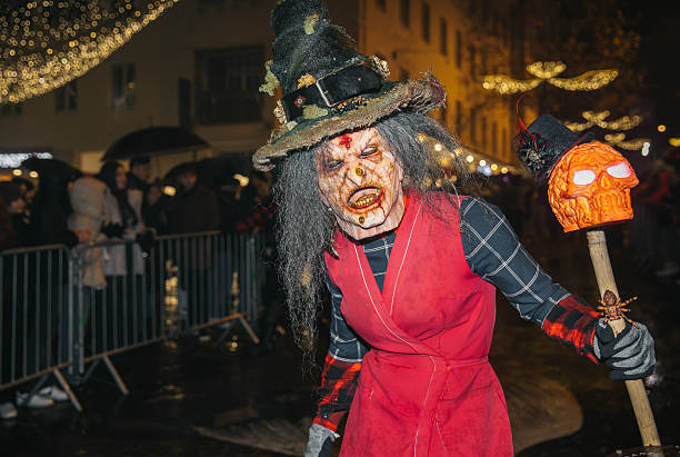Villach, Austria - December 01, 2023: Krampuslauf event showcases a costumed character with spooky makeup and a skull staff, amidst a vibrant crowd and holiday lights