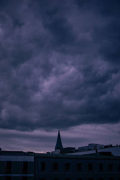 Moody dramatic sky over Munich, Germany with dark storm clouds gathering above the city skyline. A pointed church spire rises in the distance beneath the heavy clouds, creating an atmospheric and cinematic urban scene. Travel, Europe, weather, and city mood concept.