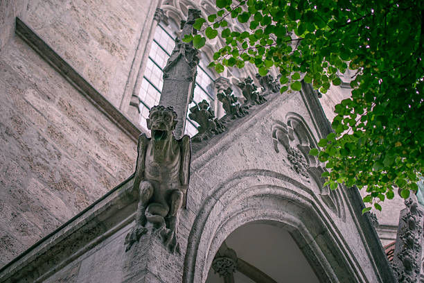 A stone gargoyle sits atop the entrance of a Gothic cathedral in Munich, Germany, framed by lush green leaves and beautiful stone carvings. The image blends dramatic medieval architecture with nature, symbolizing European history, heritage and cultural tourism.