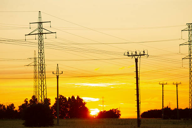 sunset over transmission corridor with towers, drone surveyor concept capturing layered pylons, vivid orange sky, long powerlines crossing frame, tree line silhouettes, energetic