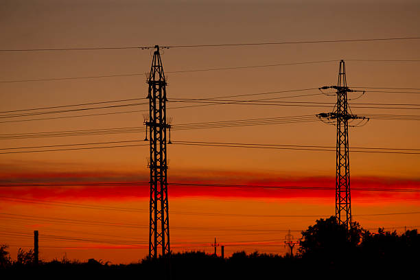 Sunset electrical towers silhouette over field, warm orange sky and layered pylons, highvoltage wires crossing frame, distant trees and calm rural horizon, moody contrast