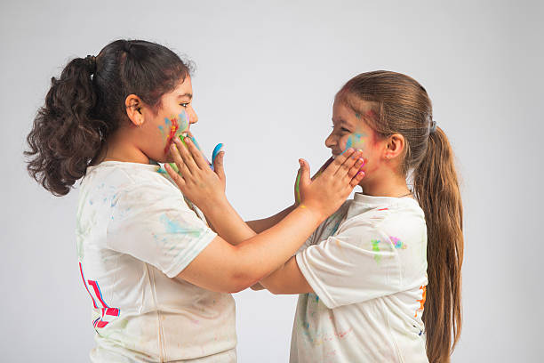 Portrait of playful sisters applying colorful powders on each other faces while celebrating Holi together on white background