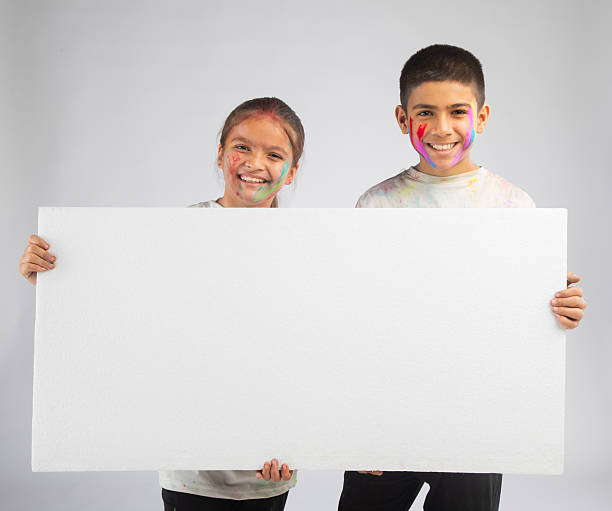 Portrait of smiling sister and brother with colorful powder on faces holding blank poster against white background