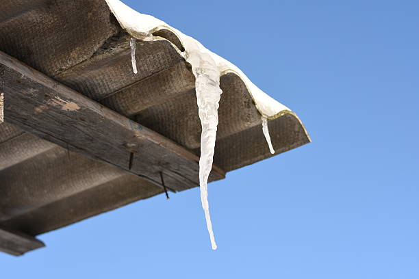 Single icicle hanging from the edge of a house roof against a clear sky. Minimal winter composition.