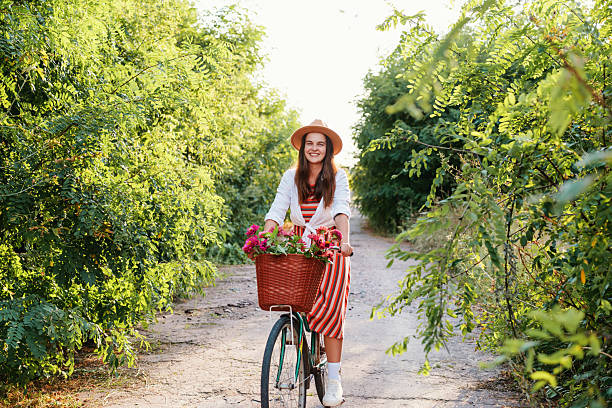 A woman with a hat rides a bicycle down a path lined with greenery. Her bicycle has a basket filled with flowers. It is daytime and sunny.